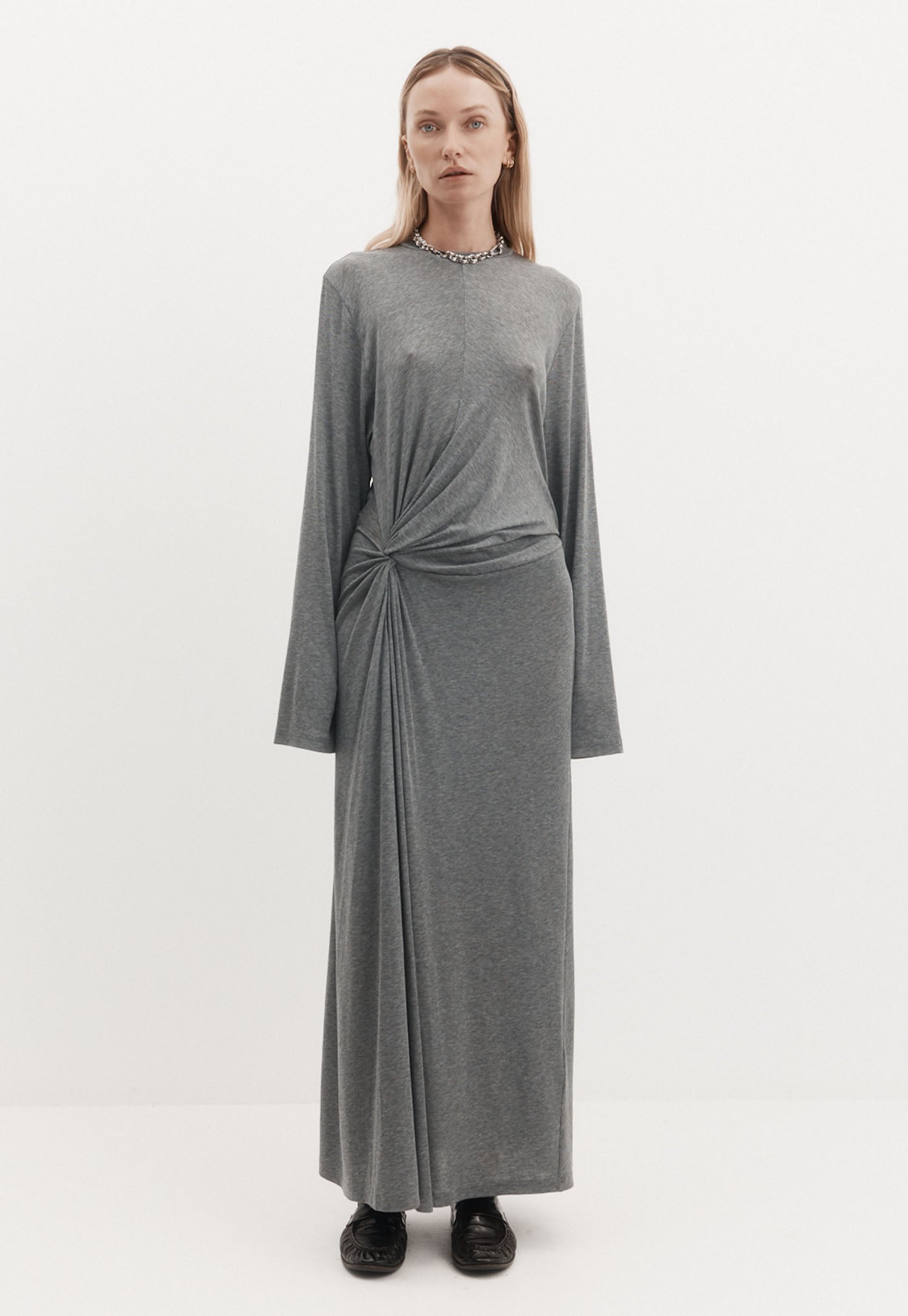A model wearing a long-sleeved gray knit maxi dress with a twisted knot detail at the waist, paired with black shoes, against a plain white studio background.